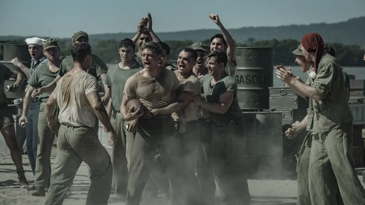 "A group of WWII-era soldiers in dusty uniforms are shouting and cheering around two men, possibly during a brawl or contest, outdoors near metal barrels and mountains in the background."
