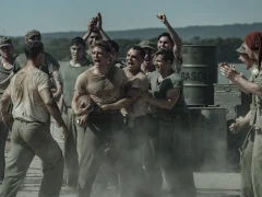 "A group of WWII-era soldiers in dusty uniforms are shouting and cheering around two men, possibly during a brawl or contest, outdoors near metal barrels and mountains in the background."